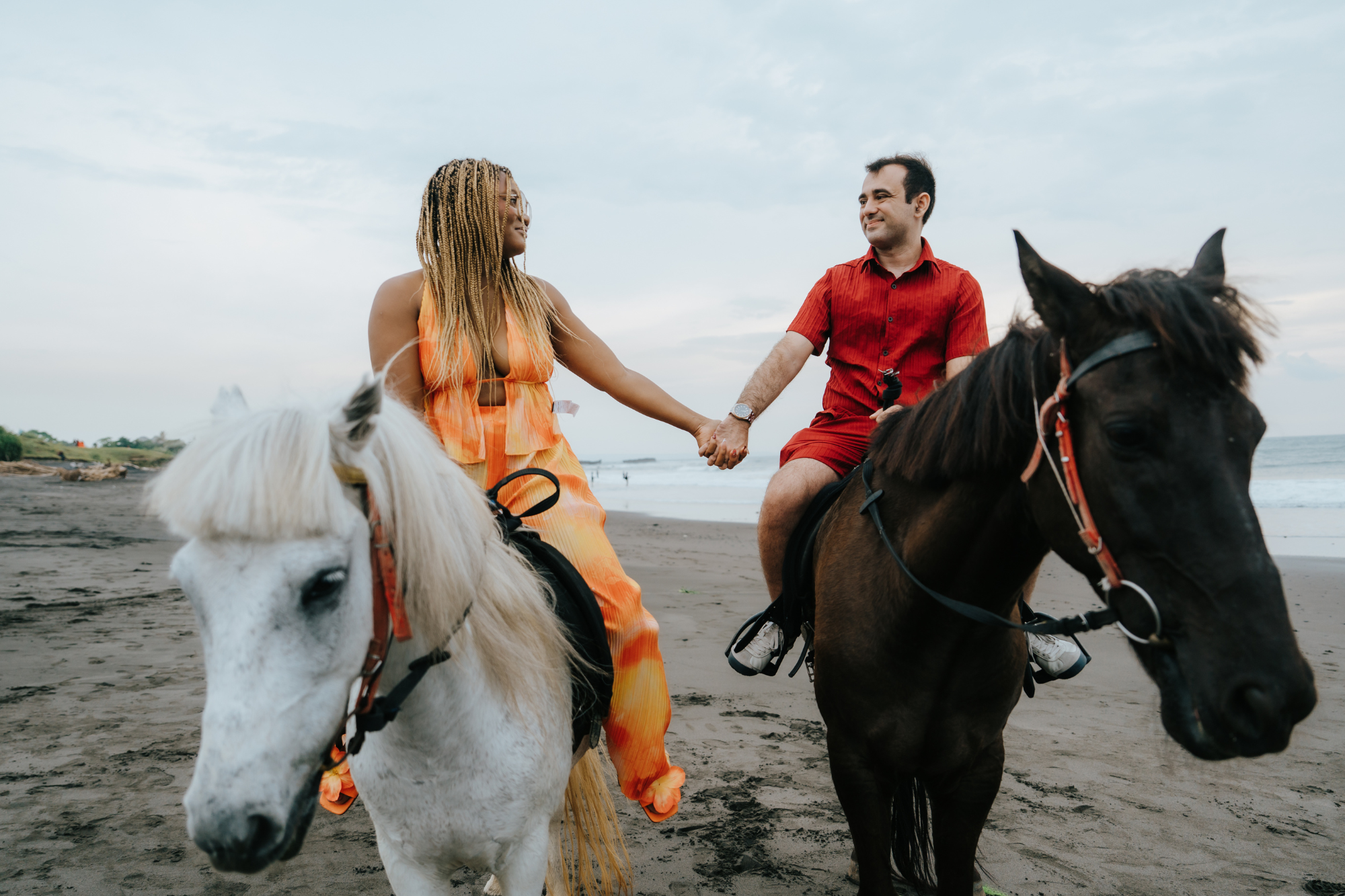 man and woman holding hands while riding horses on a bali beach