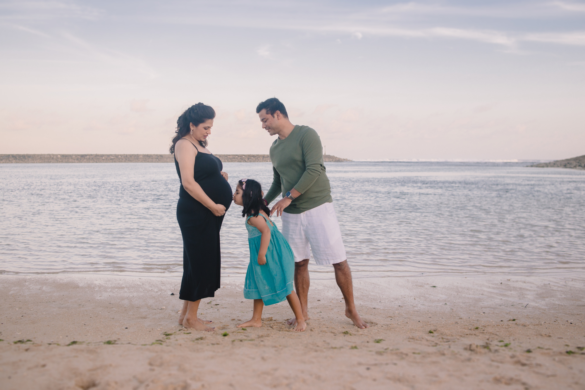 family photoshoot on bali beach: pregnant mother, daughter kissing pregnant belly and dad smiling